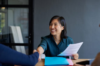 A smiling professional woman sits at a table, shaking hands with a colleague while holding a document, as another person looks on during a meeting.