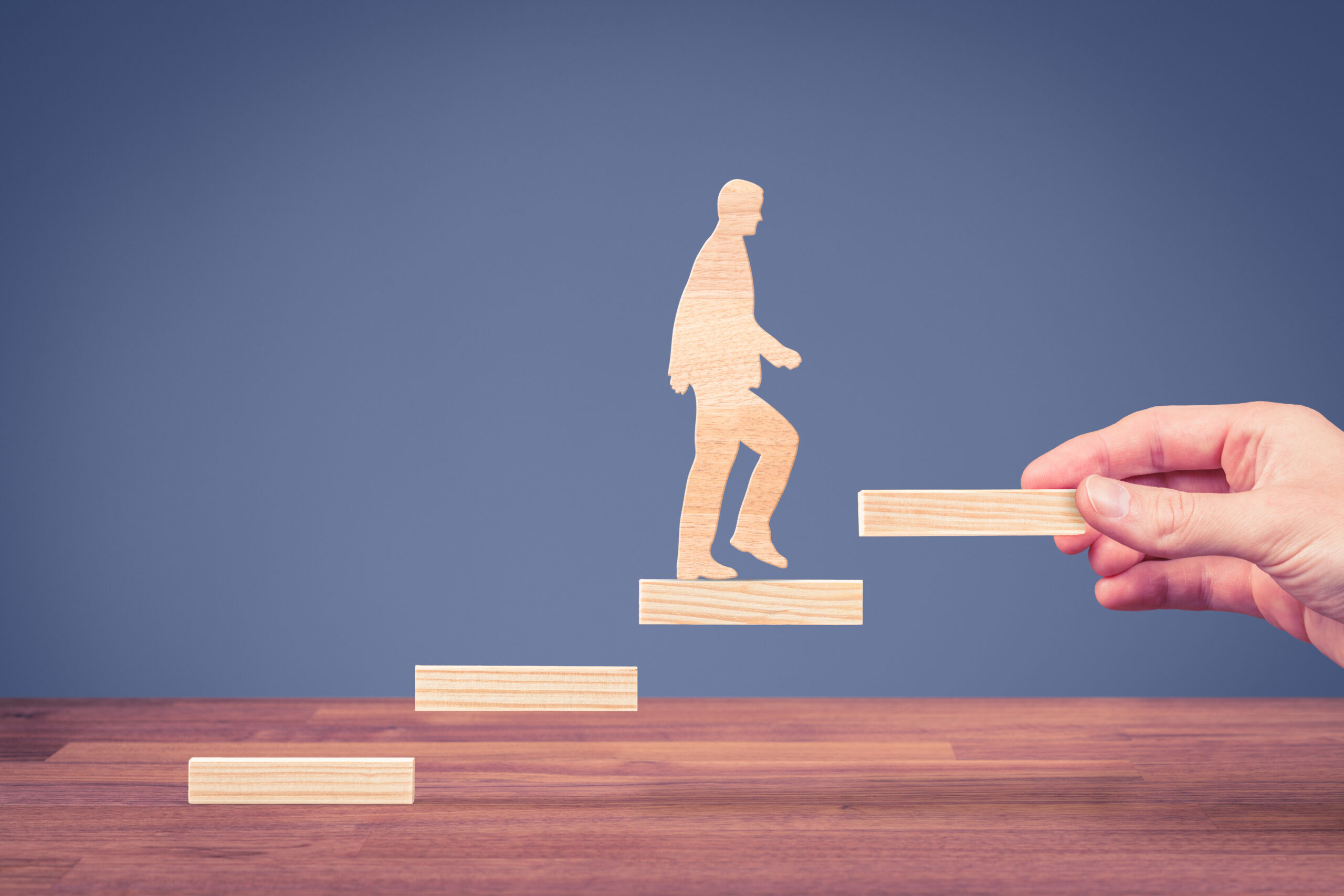 A wooden cutout of a person climbing stairs made of wooden blocks, with a hand placing the next block to help them ascend.