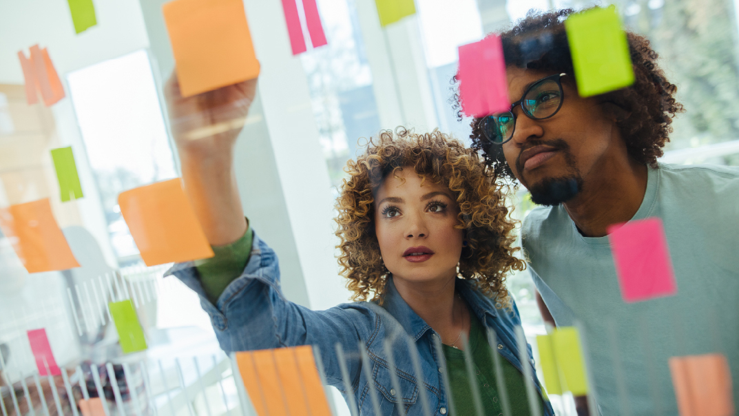 Two people stand in front of a glass wall covered with colorful sticky notes, focusing intently as they collaborate and organize ideas.