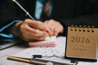 Person in a business suit writing in a small notebook at a desk, with a 2026 desk calendar in the foreground and charts on a clipboard nearby.